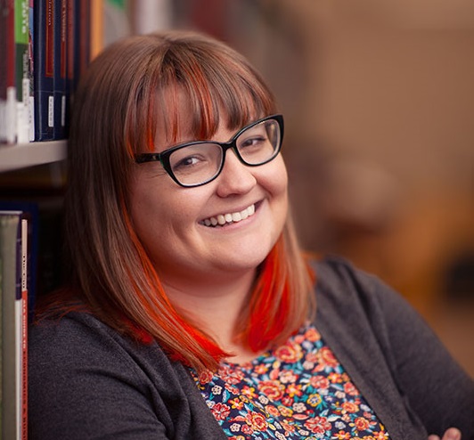 Jennifer Eltringham, a white woman with glasses, smiles next to a book shelf.